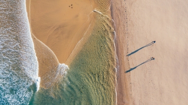 Top-down view of couple walking along sandy beach washed by Atlantic Ocean waves in Portugal casting long shadows in low sunrise light. Shot during the Camino Portuguese