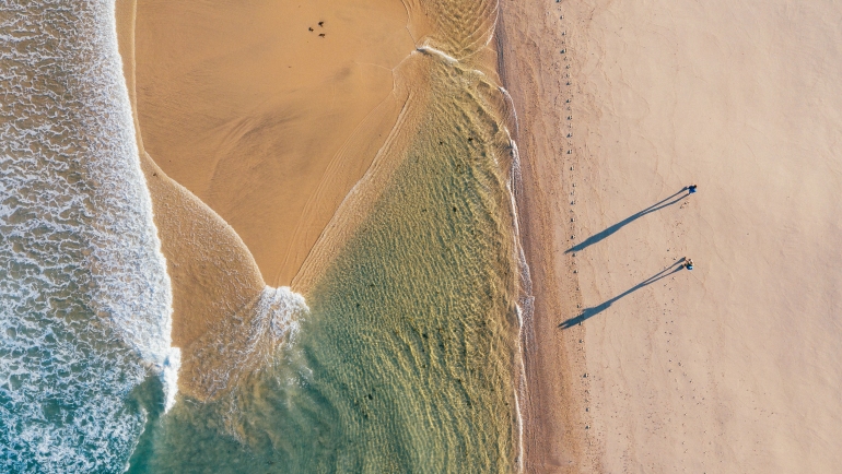 Top-down view of couple walking along sandy beach washed by Atlantic Ocean waves in Portugal casting long shadows in low sunrise light. Shot during the Camino Portuguese