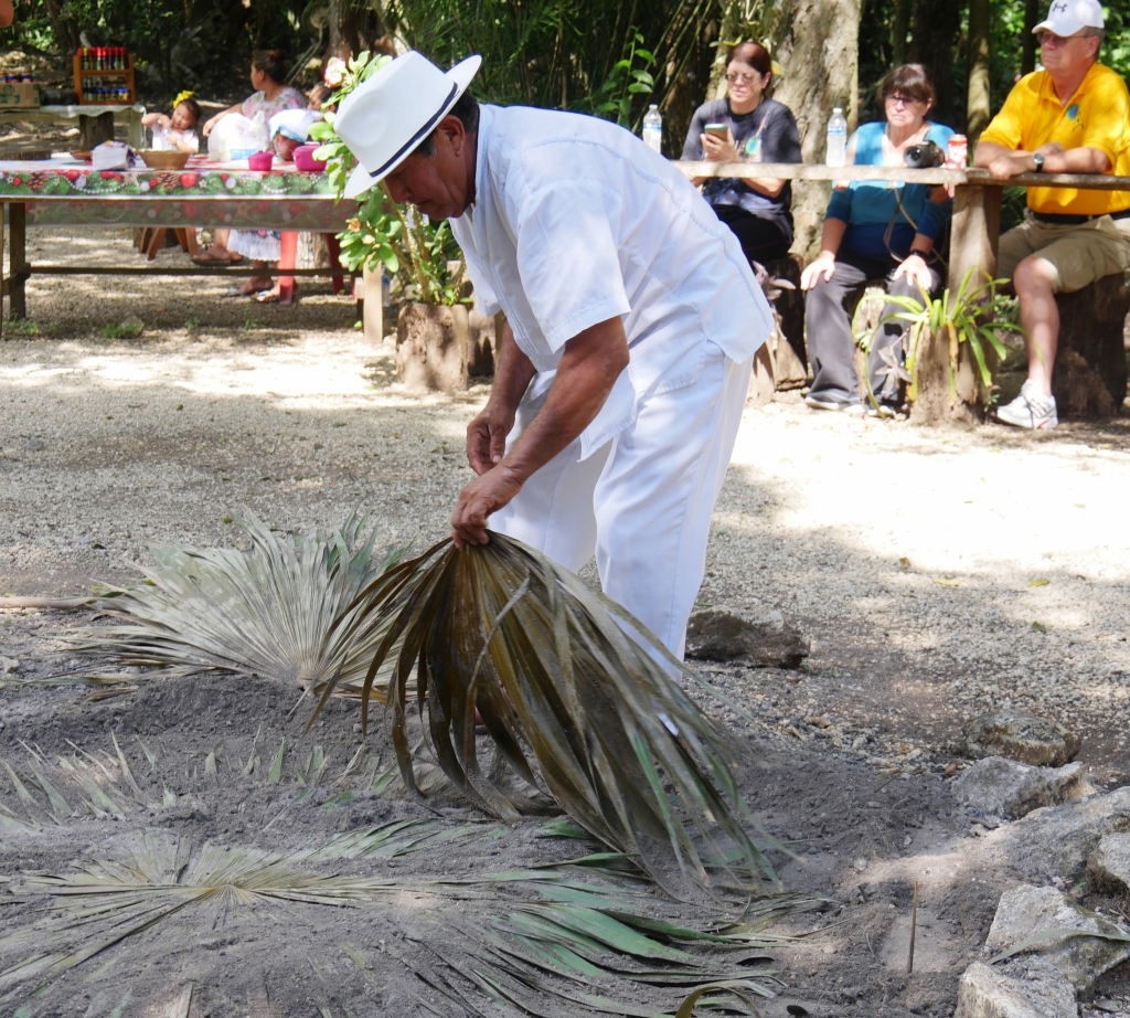 A village resident picks up the leaves covering a traditional underground stove as tourists look in in Limones village, Costa Maya