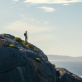 Un excursionista solitario se encuentra en una cresta de granito sobre la costa, rodeado de vegetación natural y luz del día suave. La escena destaca el escarpado terreno costero típico del sur de Australia Occidental.