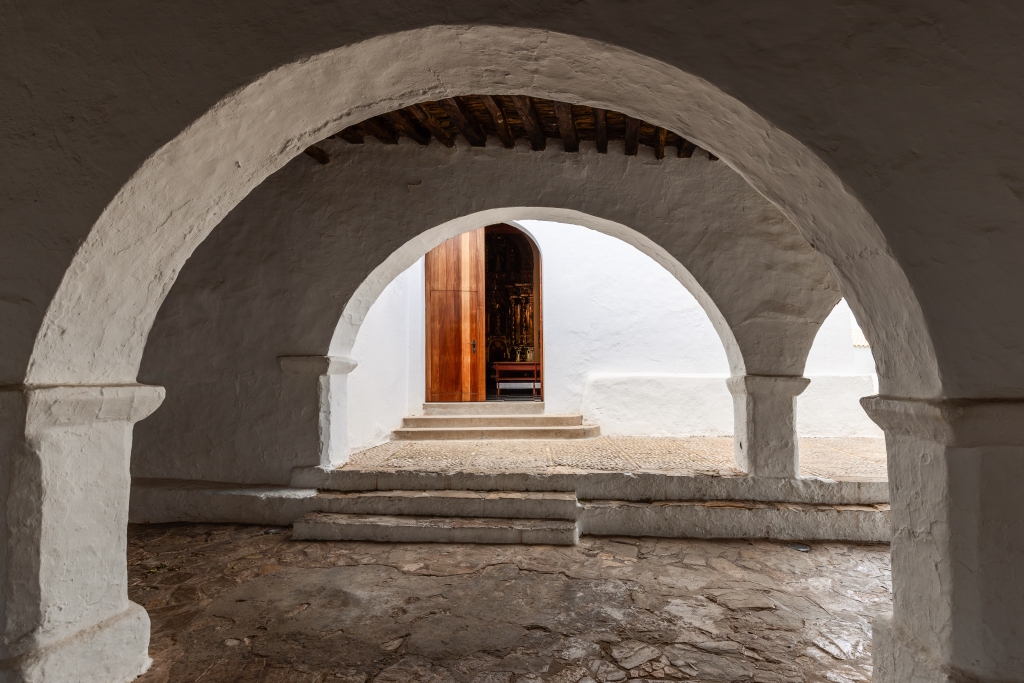 The welcoming entrance of the Puig de Missa church, framed by traditional whitewashed arches, offers a glimpse of the sacred interior through its open wooden door; the church is located in Santa Eulalia, Ibiza.