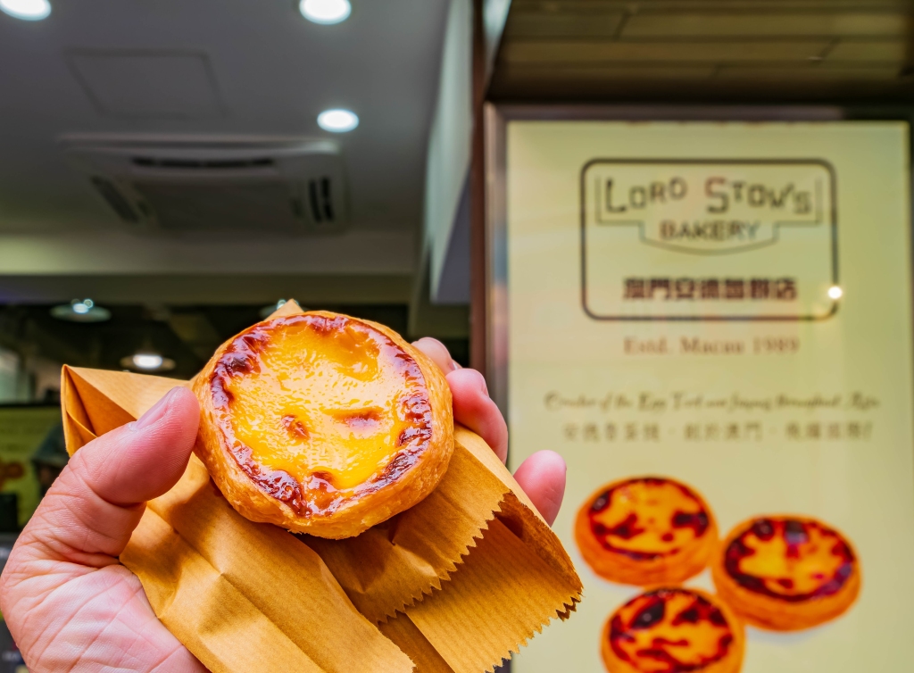 A person holds a delicious golden Portuguese egg tart wrapped in brown paper, with a sign for Lord Stow's Bakery.